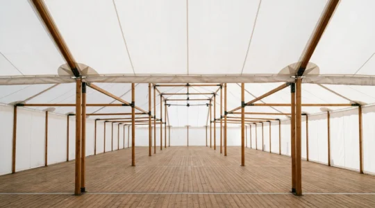 Professional editorial photograph of an empty white marquee tent interior awaiting floral transformation, with soft natural light filtering through canvas walls and visible structural beams