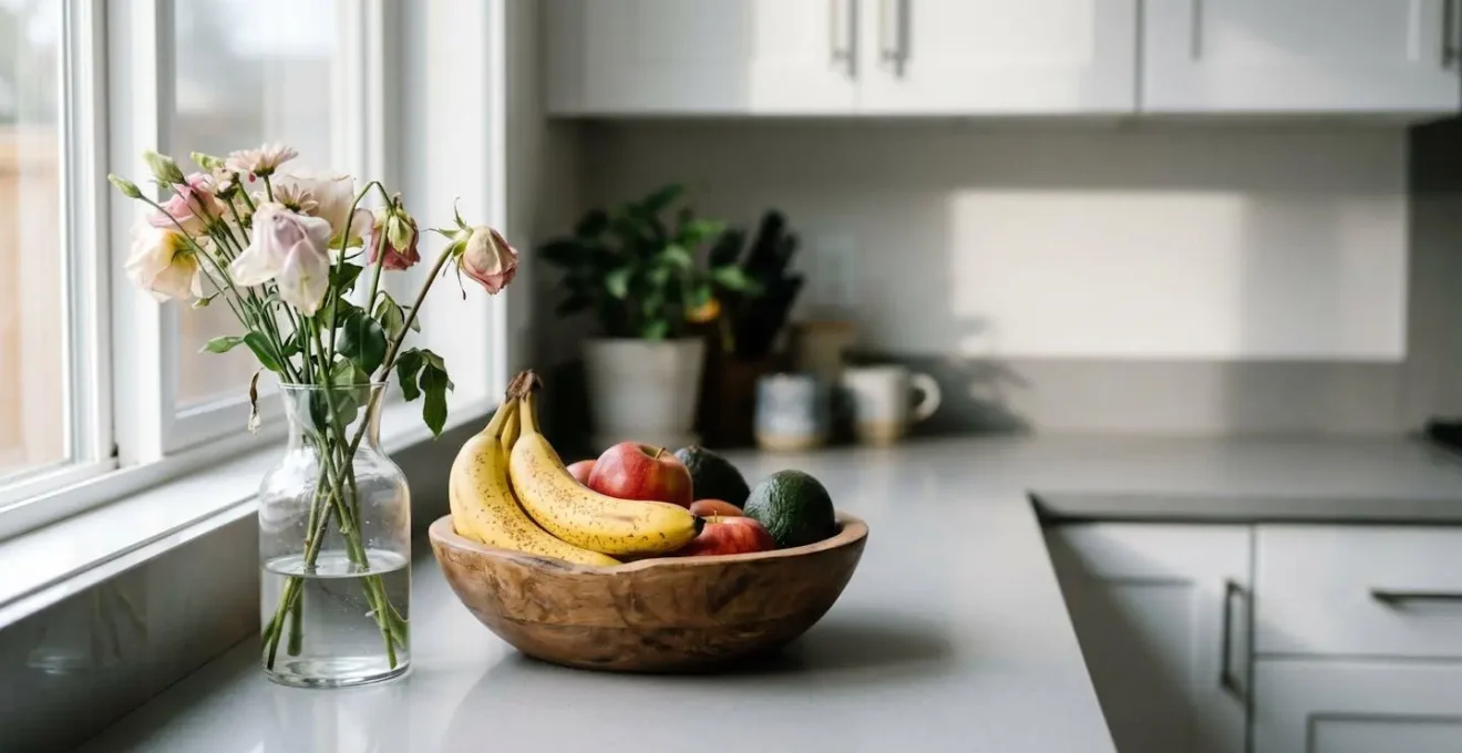 Close-up of wilting cut flowers next to a wooden fruit bowl with ripe produce on a kitchen counter