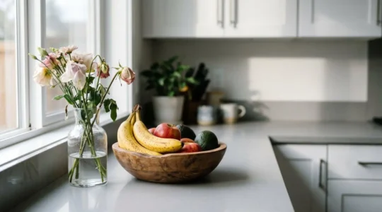 Close-up of wilting cut flowers next to a wooden fruit bowl with ripe produce on a kitchen counter